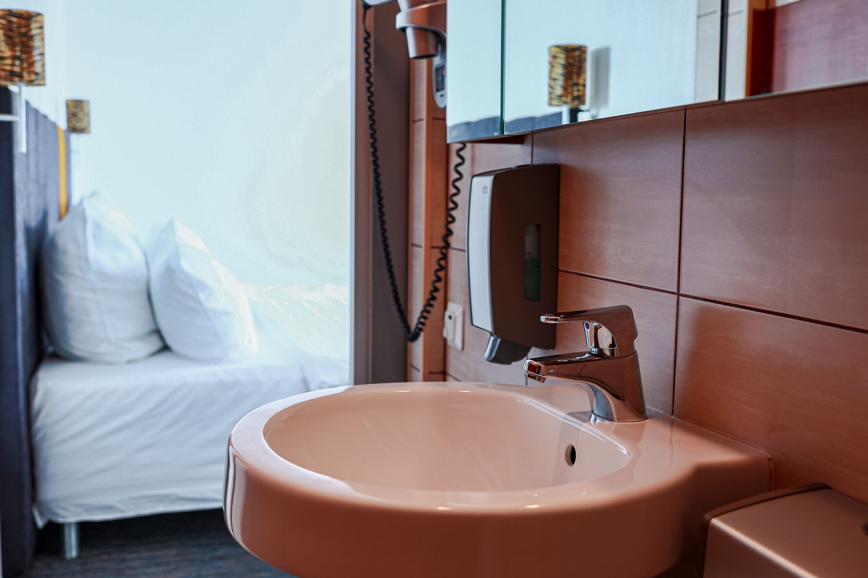 Une salle de bain avec un lavabo, un miroir et un lit en arrière-plan.