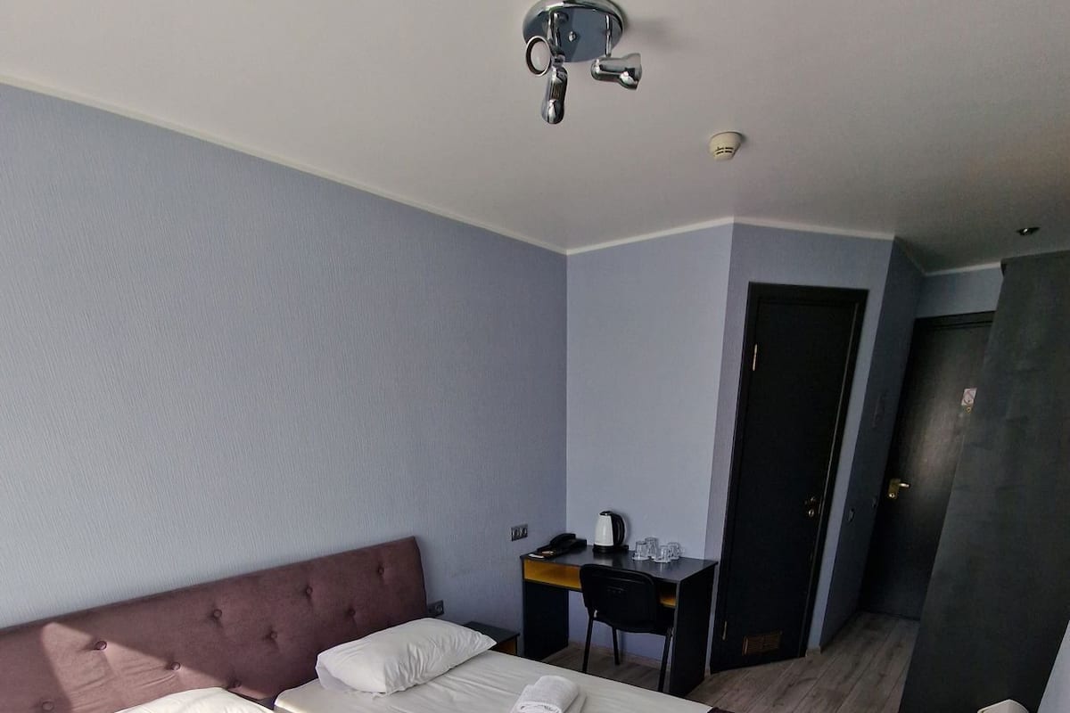 A hotel room with a double bed, a desk with a chair, a small table with a kettle, and a door.