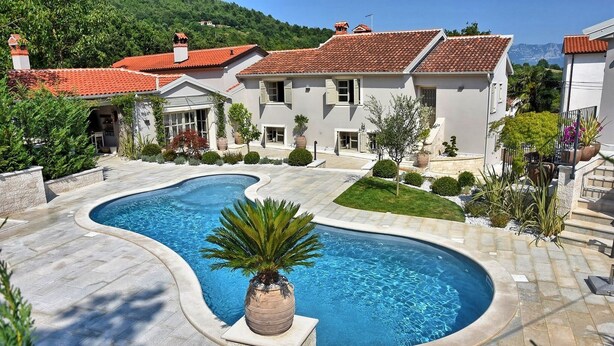 A swimming pool with a curved design, surrounded by a paved area and a house with a red-tiled roof. - Luxuriously decorated villa with designer interior, pool, sauna and wellness (Pićan)