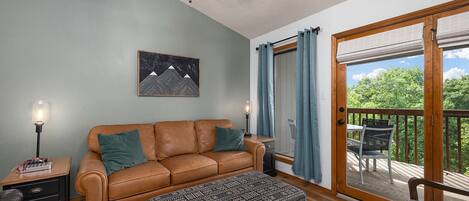 Ett mysigt vardagsrum med en brun lädersoffa, en mönstrad pall, en takfläkt och ett stort fönster med utsikt över grönområden.
