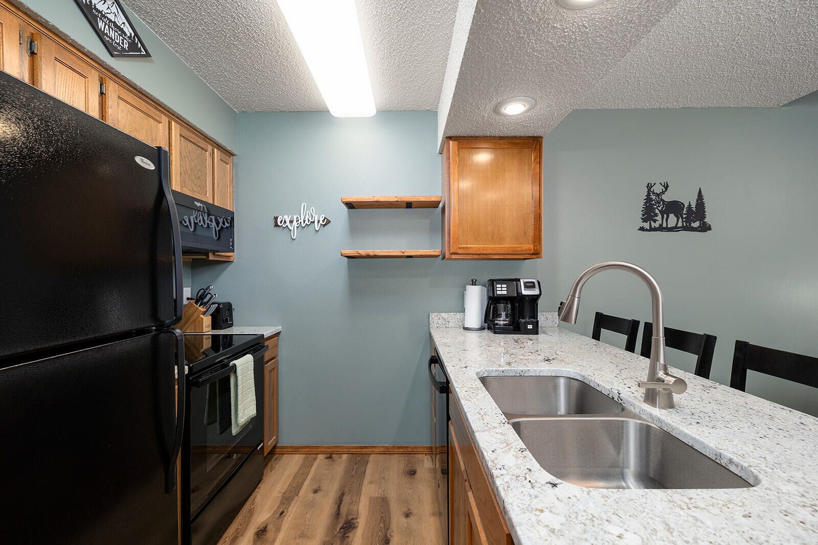 A compact kitchen with a black refrigerator, wooden cabinets, a granite countertop, a stainless steel sink, and a coffee maker.