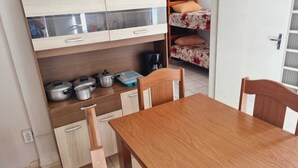A compact kitchen with wooden cabinets, a dining table, and chairs. There are pots and pans on the counter, and a bunk bed with floral bedding is visible in the background. - Apartamento completo (Guaratingueta)