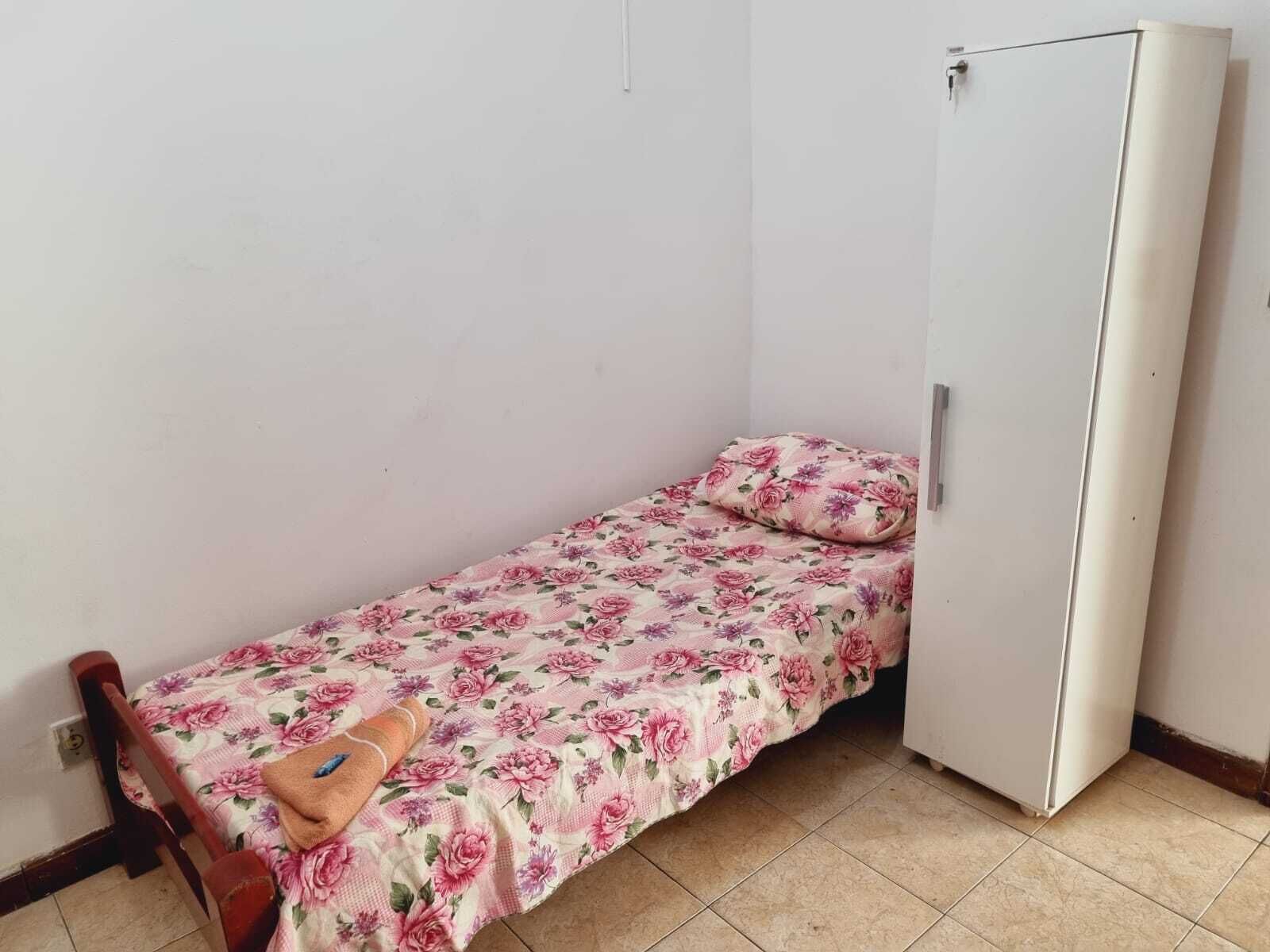 Una habitación pequeña con una cama individual, una colcha estampada, una silla de madera y un armario blanco.
