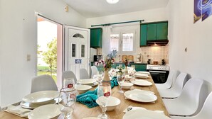 A dining area with a long table set for a meal, kitchen in the background, and a door leading outside.
