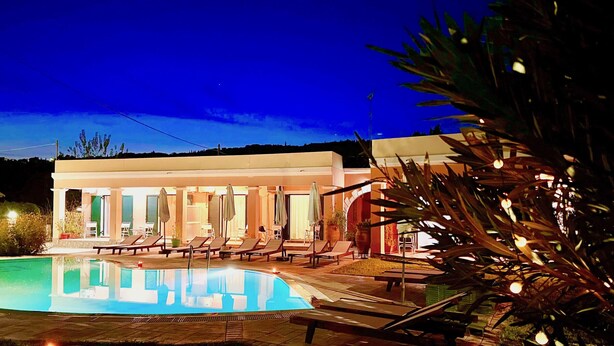 A poolside area with lounge chairs and umbrellas, a pool, and a building with multiple windows and doors.