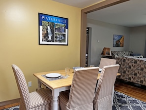 A dining area with a table set for four, chairs, a framed picture of waterfalls, and a bed in the background.