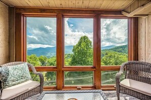 Een kamer met een groot raam met uitzicht op een bos en bergen. Er staan twee rieten stoelen met kussens.