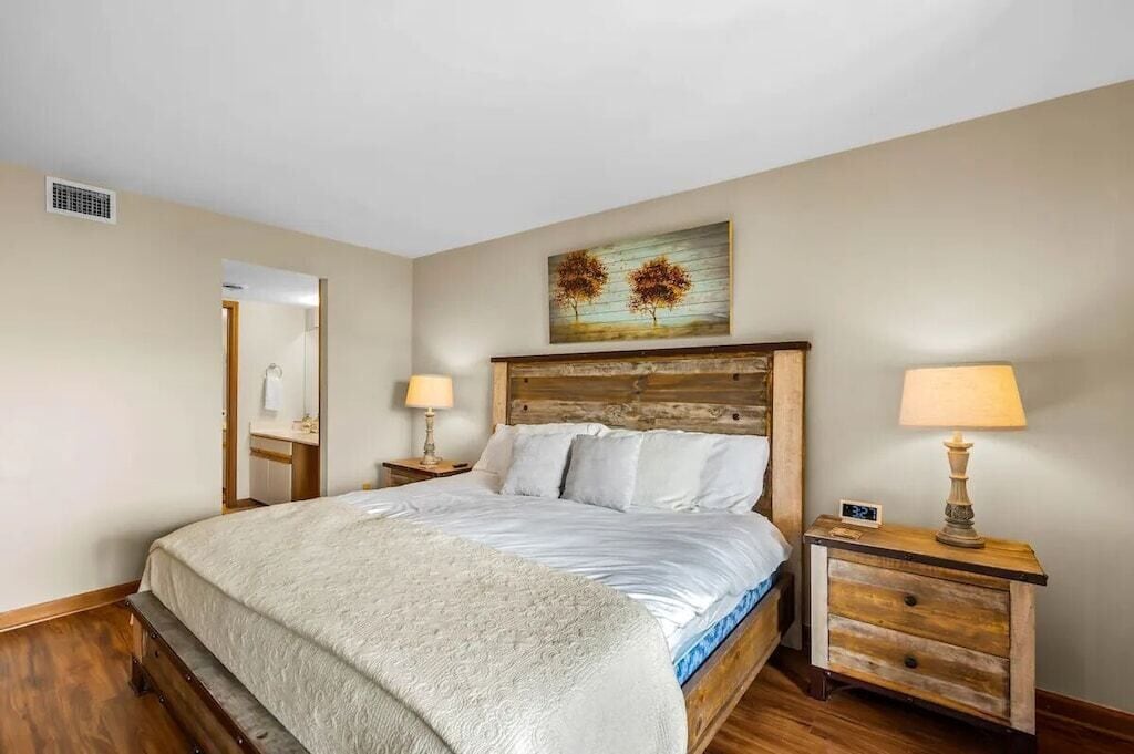 A bedroom with a wooden headboard, a bed with white linens, two bedside tables with lamps, and a painting above the headboard.
