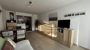 Un salon moderne avec un canapé, un téléviseur et un meuble en bois. On y trouve également un coin repas avec des chaises et une table, ainsi qu’une cuisine à l’arrière-plan.