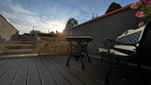 Une terrasse en bois avec une table et un banc, un mur de pierre et une voiture garée à l’arrière-plan.