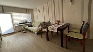 Un salon comprenant un canapé, une table à manger avec des chaises, un radiateur et une fenêtre avec des stores.