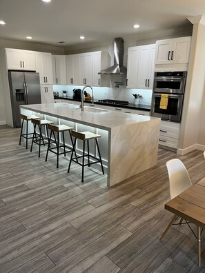 A modern kitchen with a central island, bar stools, stainless steel appliances, and wooden flooring.
