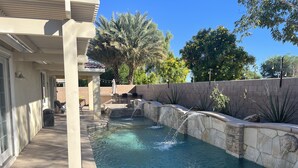 A swimming pool with a waterfall feature, surrounded by a stone wall and palm trees.