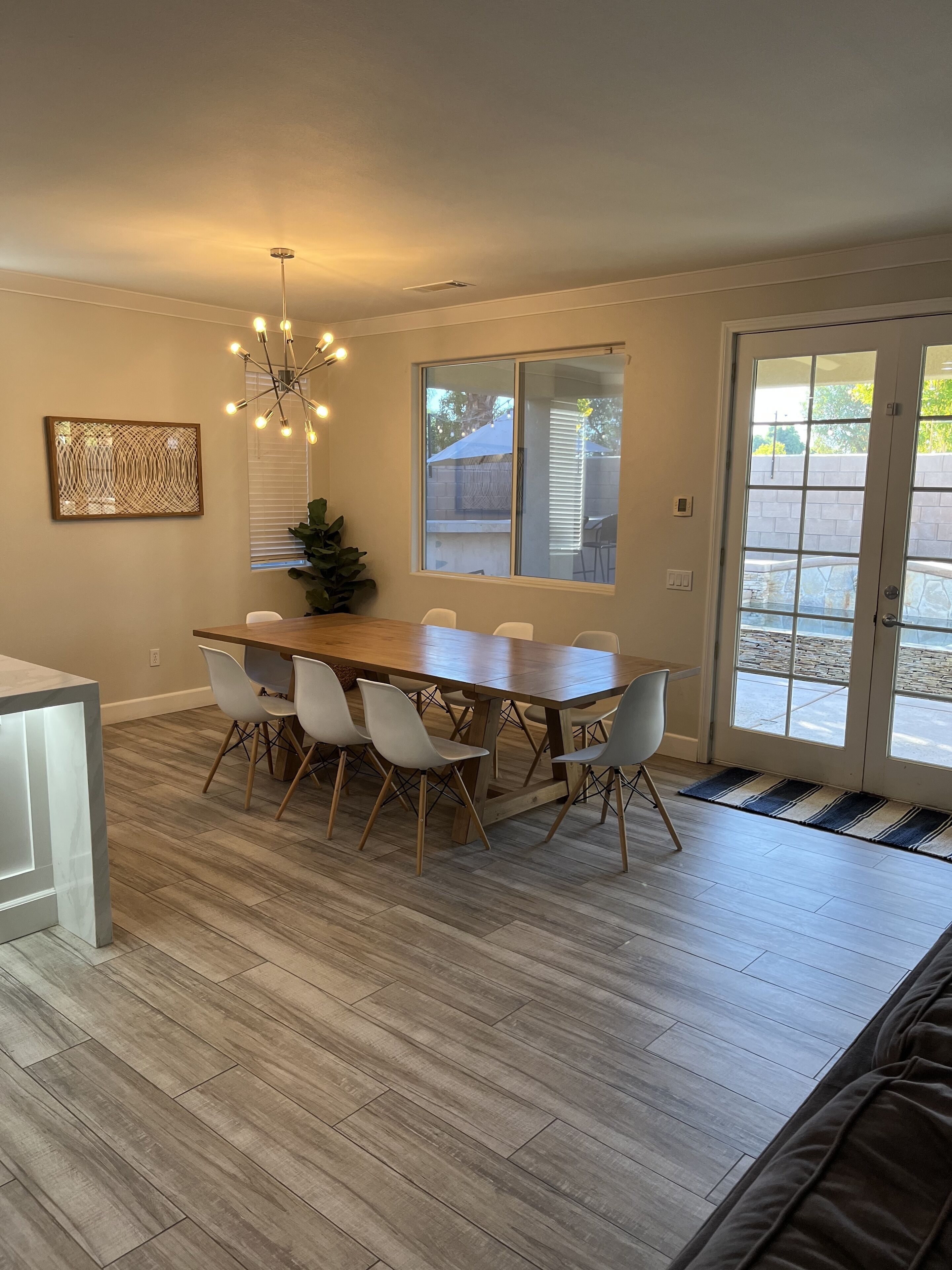 Un coin repas avec une table en bois et des chaises blanches, un lustre et une vue à travers une porte vitrée.