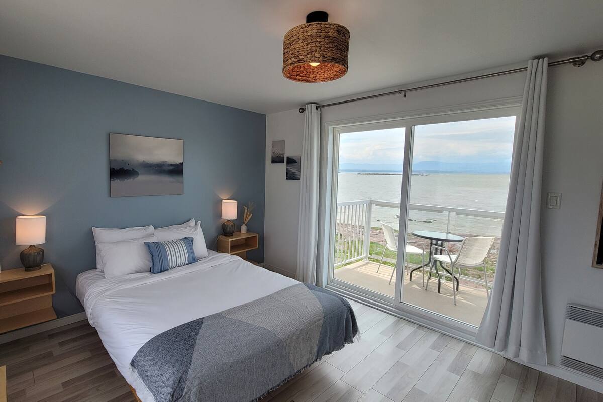 A bedroom with a large bed, bedside lamps, a painting, and a view of the sea through a sliding glass door.
