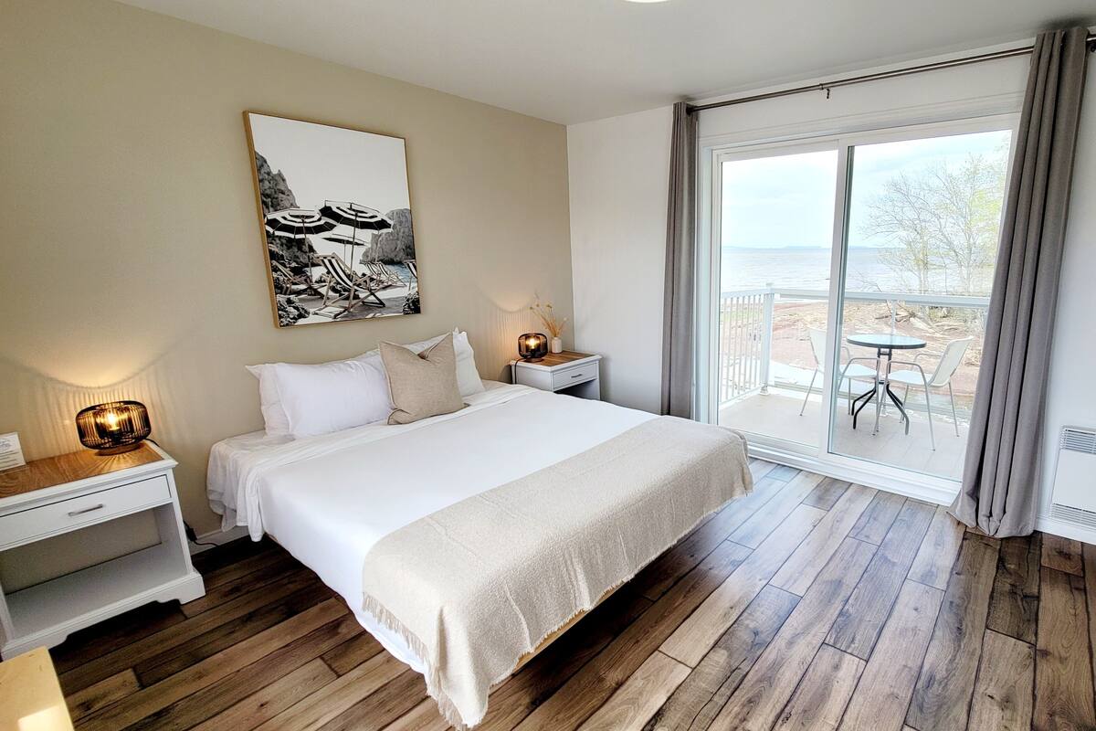 A bedroom with a large bed, a nightstand with a lamp, a bedside table, and a view of the balcony with a table and chairs.