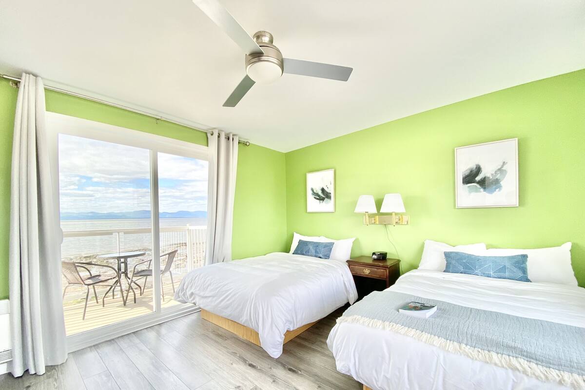 A hotel room with two beds, a ceiling fan, a balcony with a table and chairs, and artwork on the walls.