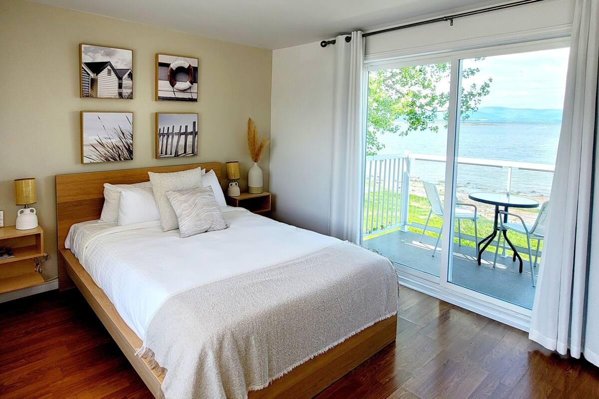 A bedroom with a wooden bed, white bedding, a nightstand with a lamp, a small shelf with a vase, and a view of a balcony with a table and chairs.