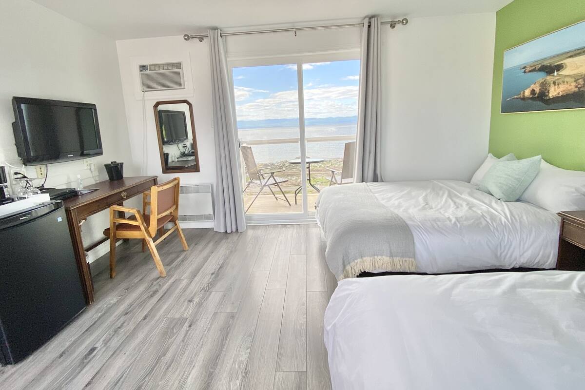 A hotel room with a bed, a desk with a chair, a TV, and a view of the sea.