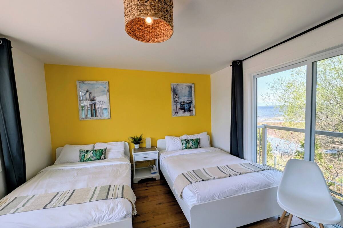 A room with two beds, a small table, and a chair. There are two paintings on the wall, a window with a view of the outdoors, and a hanging light fixture.