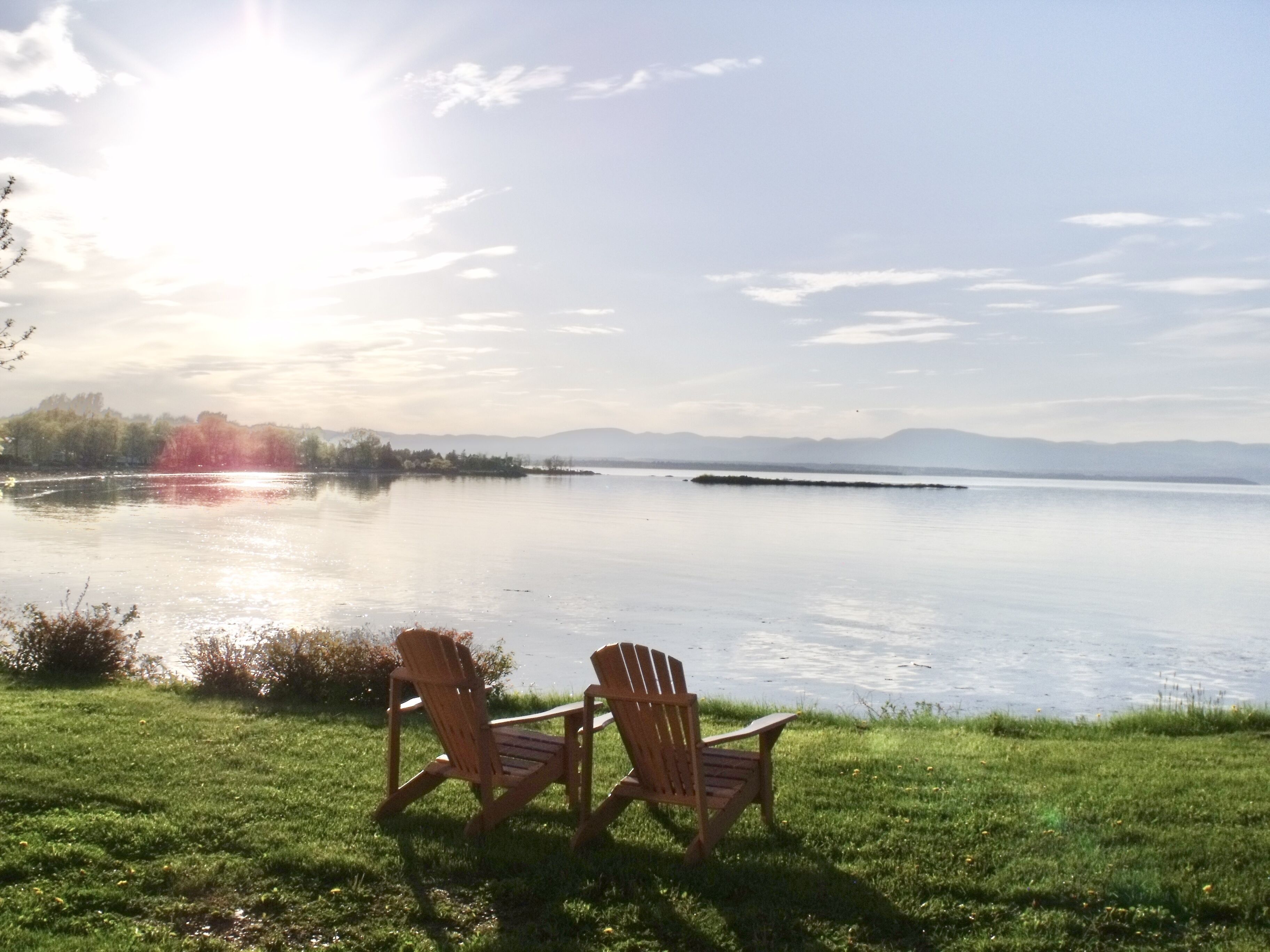 Deux fauteuils Adirondack sont placés face à un lac paisible, avec une rive et des montagnes visibles au loin.