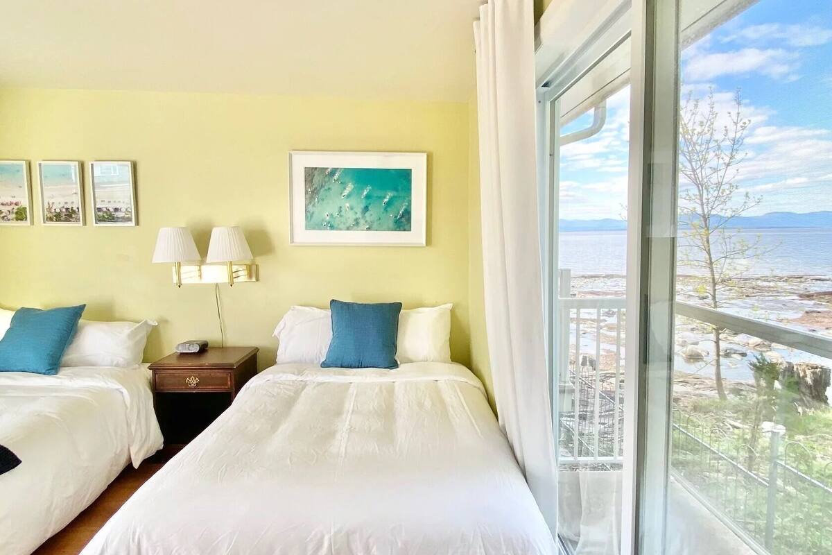 A hotel room with two beds, a nightstand, a lamp, and a large window with a view of a beach and mountains.