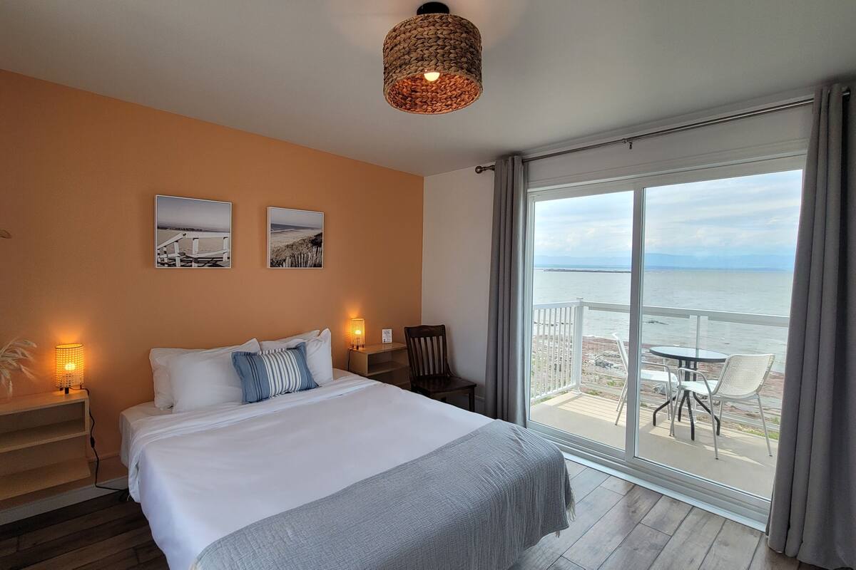A hotel room with a bed, a nightstand, a lamp, a chair, and a balcony with a view of the sea.