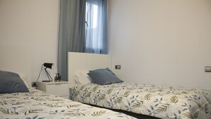 A bedroom with two beds, a nightstand with a lamp, and a window with sheer curtains.