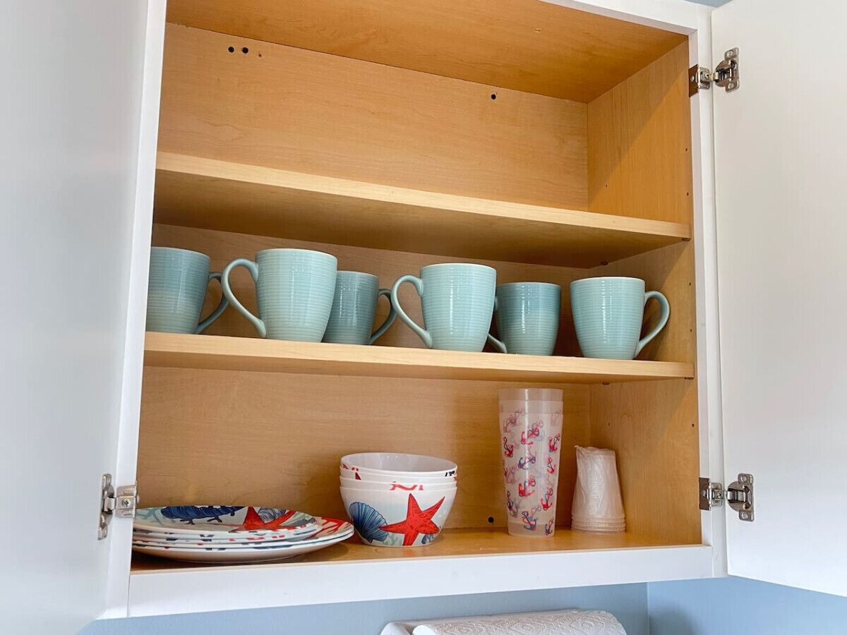 A cabinet with three shelves; the top shelf holds multiple teal mugs, the middle shelf has a bowl with a red star design, and the bottom shelf contains a stack of pink cups and a clear plastic container with a pattern.