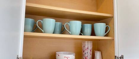 A cabinet with three shelves; the top shelf holds multiple teal mugs, the middle shelf has a bowl with a red star design, and the bottom shelf contains a stack of pink cups and a clear plastic container with a pattern.