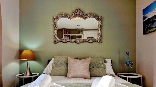 A neatly made bed with rolled towels, a large ornate mirror, and a lamp on a bedside table. - Bowness Hideaway (Windermere)