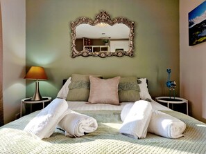 Un lit bien fait, avec des serviettes pliées, un grand miroir décoratif et une lampe sur une table de chevet.