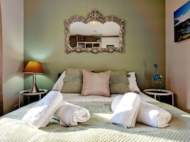 Cama bem arrumada com toalhas dobradas, um espelho grande e decorativo, e um abajur em uma mesa de cabeceira.