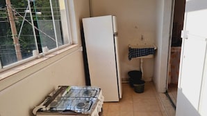 Una cocina pequeña y estrecha con una ventana, un refrigerador blanco, una estufa a gas, un lavamanos y un balde.