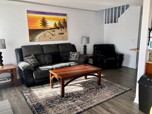 A living room with a black leather sofa, a wooden coffee table, a patterned rug, a painting of a beach scene, and a staircase.