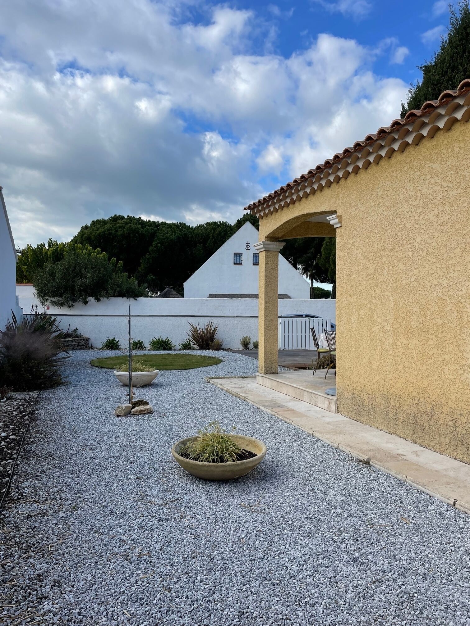 Une cour avec un chemin de gravier, une plante en pot et un bâtiment blanc doté d’un toit en tuiles.