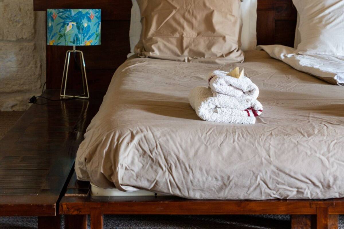 A neatly made bed with beige bedding and pillows, a wooden nightstand with a lamp, and a stone wall in the background.