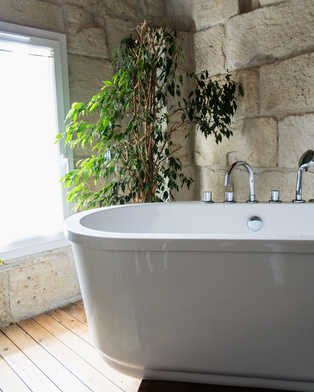 A modern bathroom with a freestanding bathtub, a stone wall, and a window.