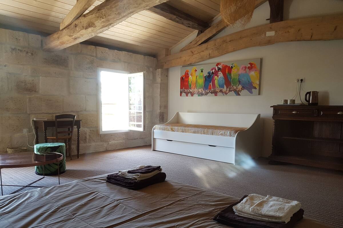 A room with a stone wall, wooden beams, a white bed, a painting of colorful parrots, a green chair, and a wooden cabinet.