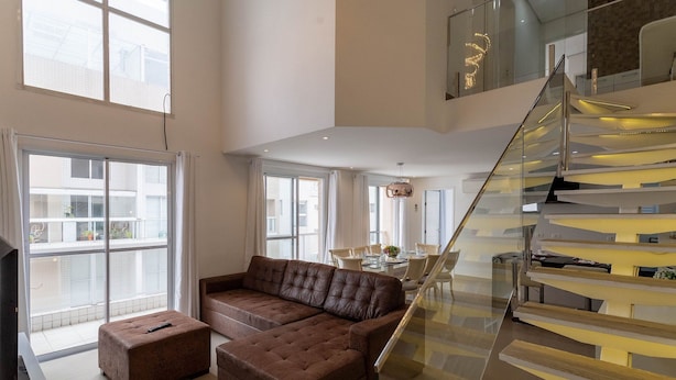 Un vaste espace de vie comprenant un grand canapé d’angle marron, un escalier en verre et un coin repas avec une table et des chaises.