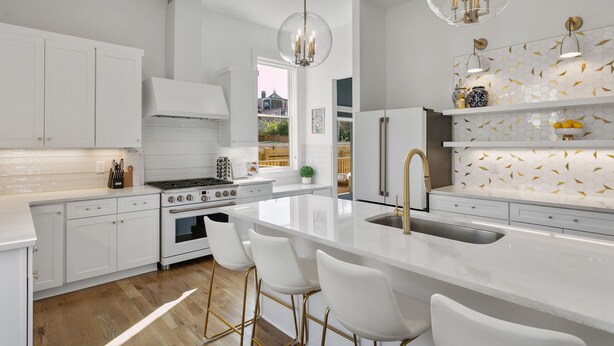 A modern kitchen with white cabinetry, a central island with bar stools, a stainless steel sink, and a decorative backsplash.