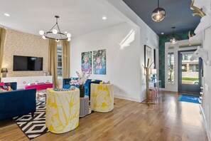 A modern living room with a large flat-screen TV, blue sofas, a patterned ottoman, and a chandelier.