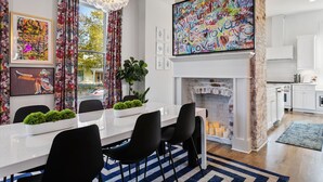 A modern dining area with a white table, black chairs, and a chandelier. A fireplace with a brick wall and a large 'LOVE' painting is visible.