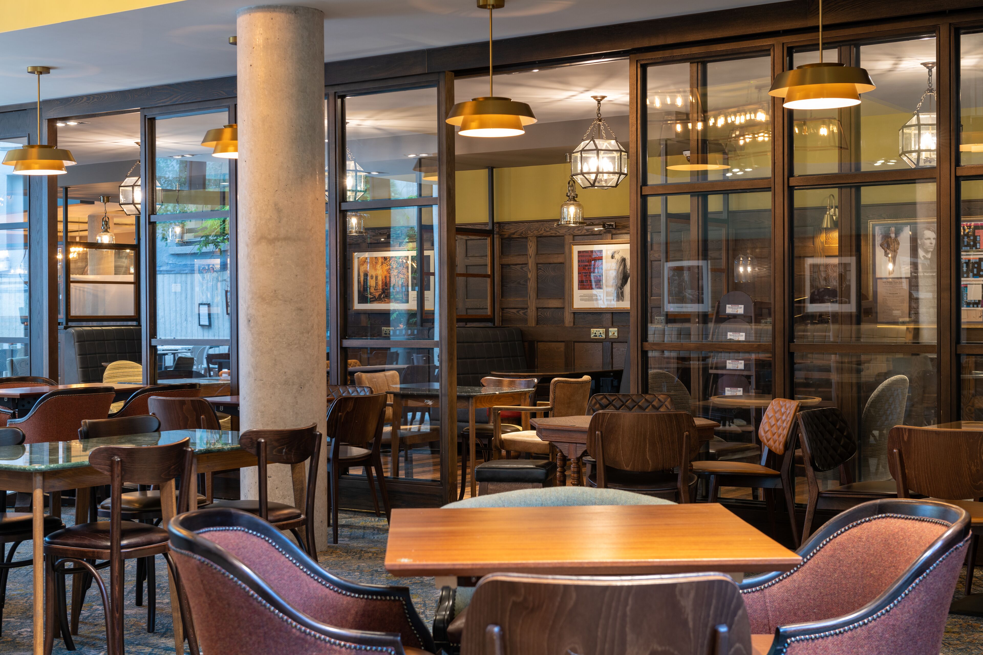 A restaurant with wooden tables and chairs, hanging pendant lights, and large windows with artwork.