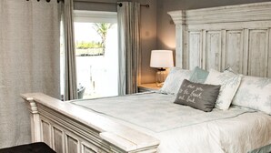 A bedroom with a large bed, a nightstand with a lamp, and a view of the outdoors.