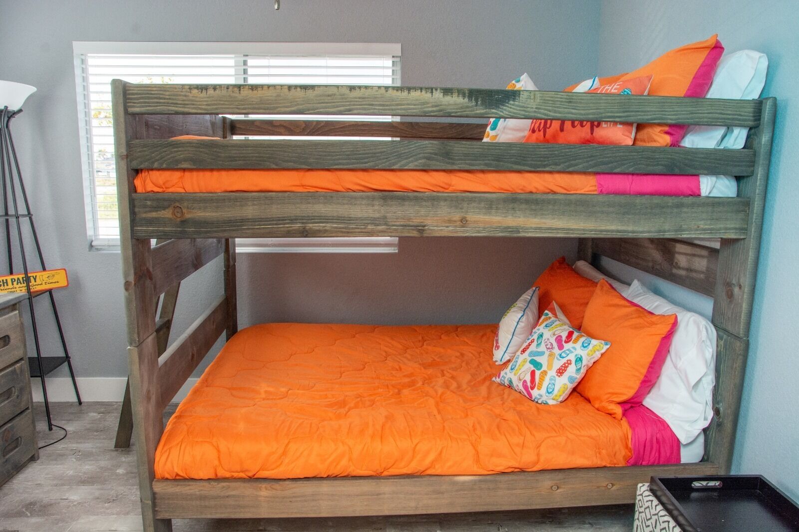 Ein Stockbett mit orangefarbener Bettdecke und Kissen, ein Holzkopfbrett und ein Fenster mit Jalousien.