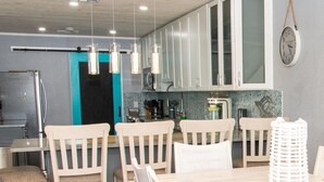 A modern kitchen with white cabinets, a teal door, hanging pendant lights, and a clock on the wall.