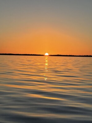 Un coucher de soleil sur une étendue d’eau calme, avec la silhouette d’arbres à l’horizon.