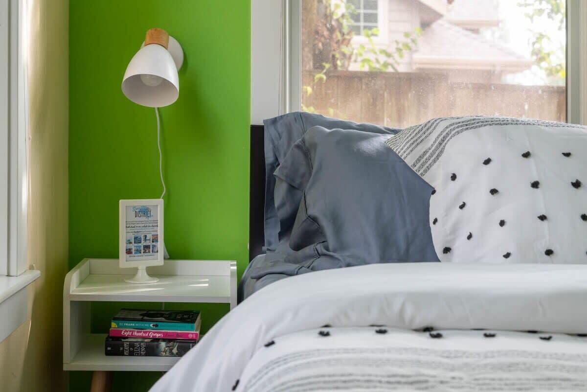 Une chambre avec un mur vert, une table de chevet blanche avec une lampe, un lit recouvert d’une couverture à pois blancs et noirs, et une fenêtre donnant sur un paysage verdoyant.