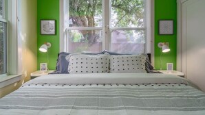 A bedroom with a large bed, green walls, two bedside tables with lamps, and a window with a view of trees. - #StayInMyDistrict Lake Oswego Honey Bee Cottage (Lake Oswego)